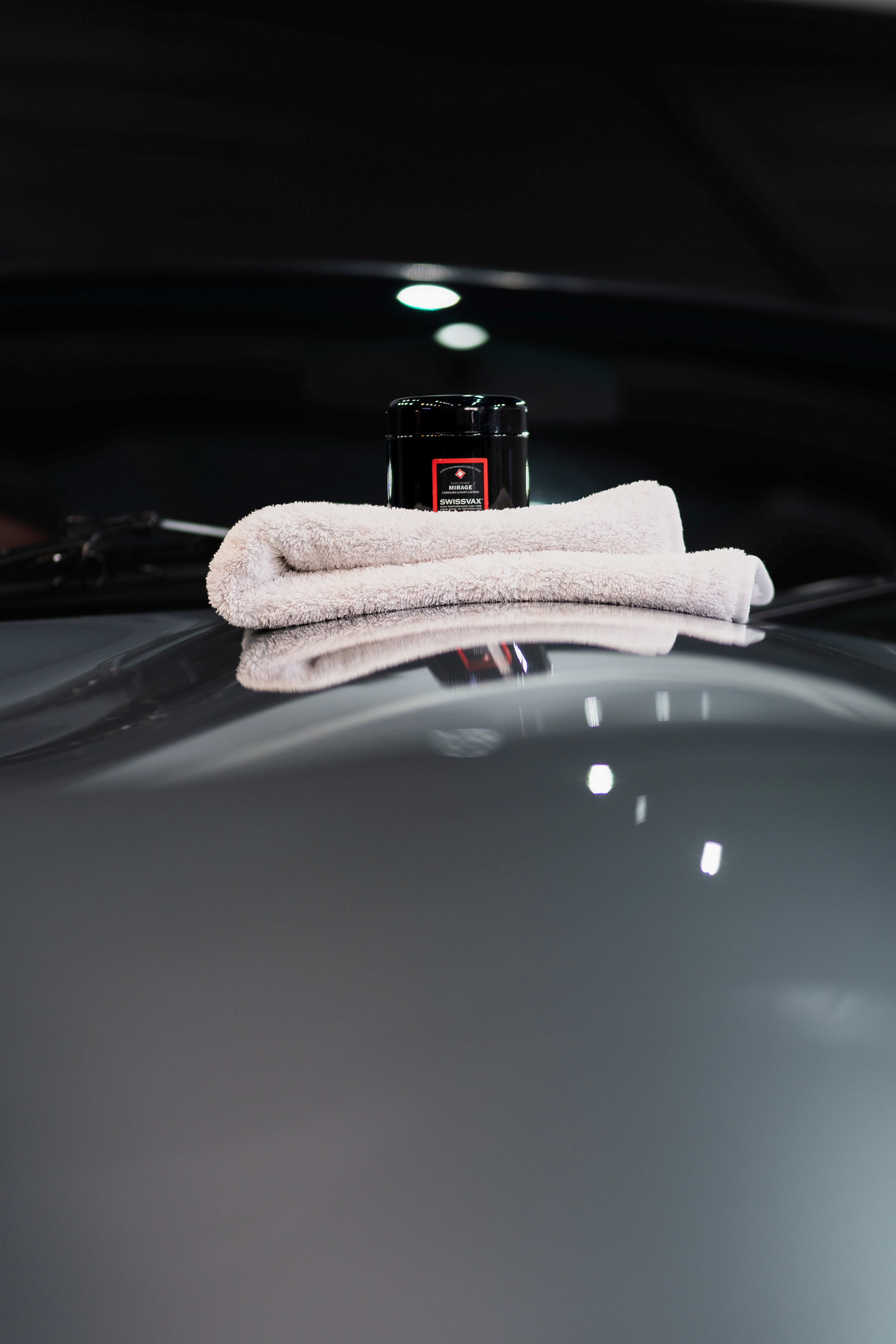 Cire Swissvax Mirage sur microfibre, posée sur le toit d'une carrosserie grise sous éclairages en spots d'atelier